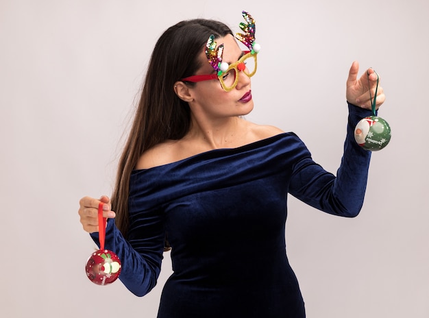 Foto gratuita pensando joven hermosa vestida con vestido azul y gafas de navidad sosteniendo y mirando bolas de navidad aisladas sobre fondo blanco.