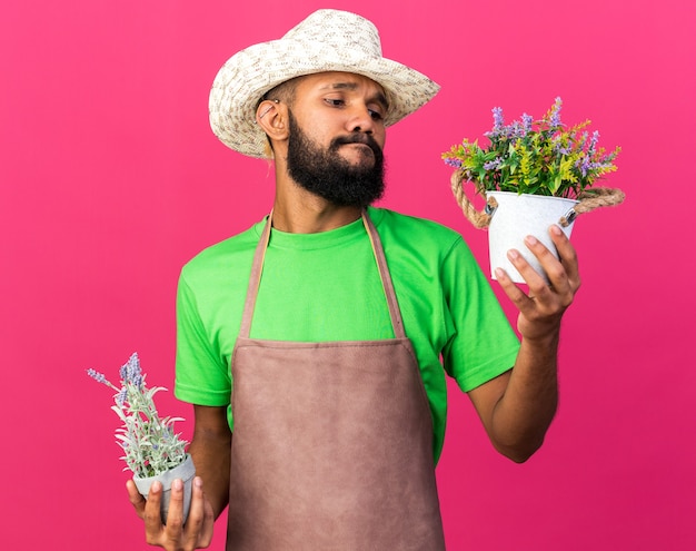 Pensando joven jardinero chico afroamericano con sombrero de jardinería sosteniendo y mirando flores en maceta