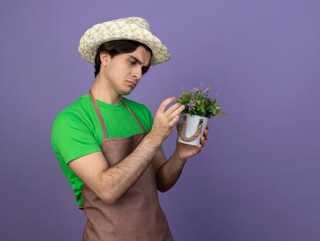 Pensando joven jardinero en uniforme vistiendo sombrero de jardinería sosteniendo y mirando flor en maceta