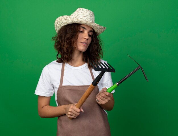 Pensando joven jardinero en uniforme vistiendo sombrero de jardinería sosteniendo y mirando rastrillo con rastrillo azada