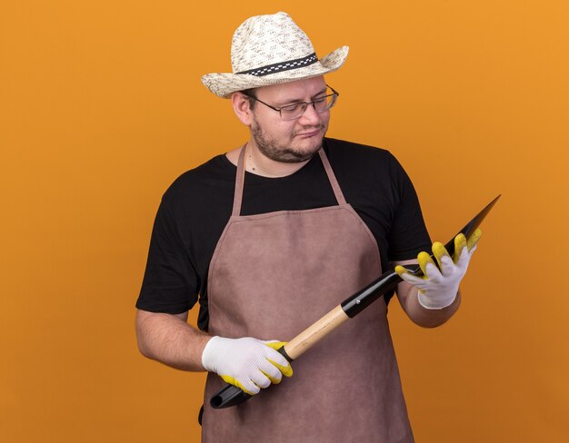 Pensando joven jardinero vistiendo gorro y guantes de jardinería sosteniendo y mirando la pala aislado en la pared naranja