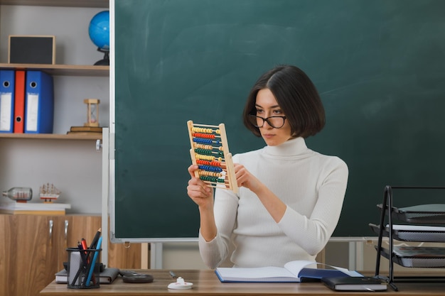 Pensando en una joven maestra con anteojos sosteniendo y mirando a un ábaco sentada en un escritorio con herramientas escolares en el aula