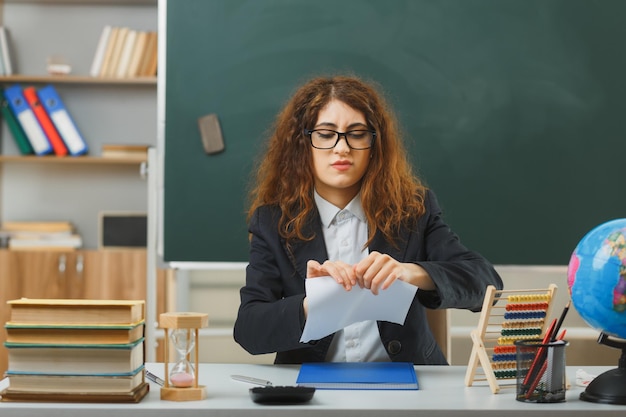 Pensando en una joven maestra con gafas, papel rasgado sentado en el escritorio con herramientas escolares en el aula