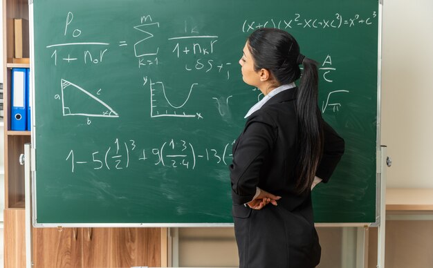 Pensando joven maestra de pie delante de la pizarra poniendo la mano en la cadera en el aula