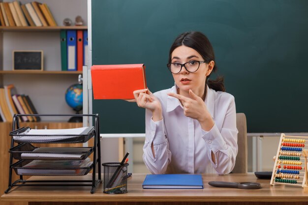 pensando en una joven maestra sosteniendo y señalando un libro sentado en el escritorio con herramientas escolares en el aula