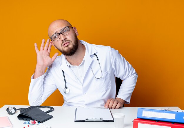 Pensando en el joven médico varón calvo vistiendo bata médica y un estetoscopio en gafas sentado