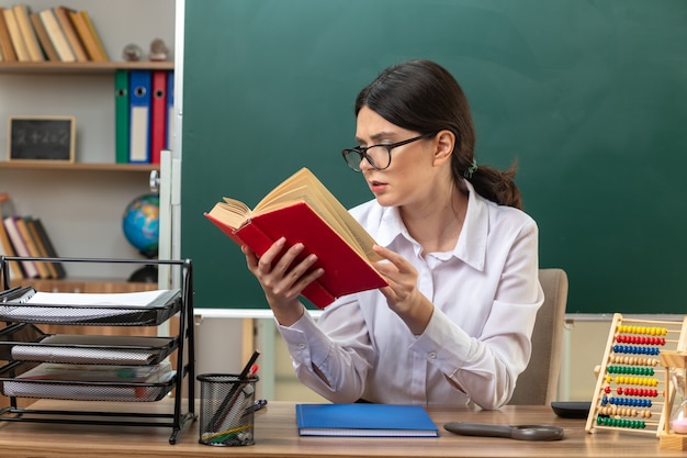 Pensando en la joven profesora con gafas leyendo un libro sentado a la mesa con herramientas escolares en el aula