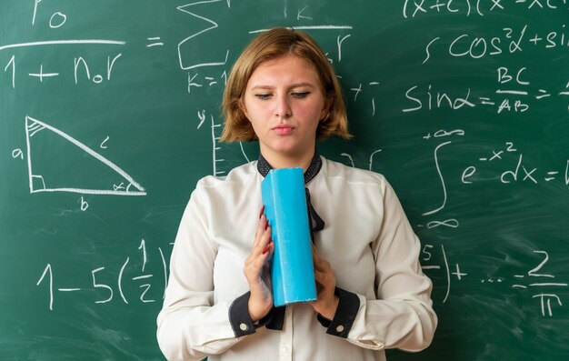 Pensando en la joven profesora de pie delante de la pizarra sosteniendo y mirando el libro en el aula