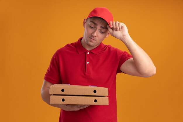 Pensando en el joven repartidor vestido con uniforme con gorra sosteniendo y mirando cajas de pizza aisladas en la pared naranja