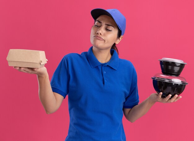 Pensando en la joven repartidora vestida con uniforme con gorra sosteniendo y mirando contenedores de alimentos aislados en la pared rosa
