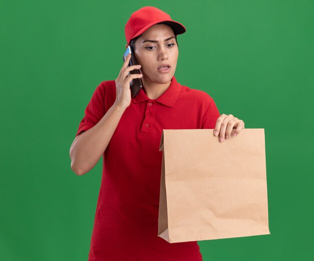 Pensando en la joven repartidora vestida con uniforme y gorra sosteniendo y mirando el paquete de comida de papel habla por teléfono aislado en la pared verde