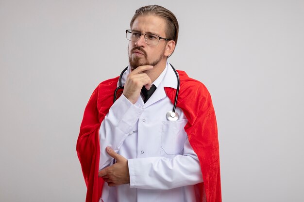 Pensando en el joven superhéroe chico mirando de lado vistiendo bata médica con estetoscopio y gafas agarró la barbilla aislado sobre fondo blanco.