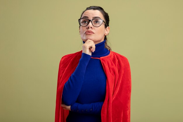 Pensando en la joven superhéroe mirando hacia arriba con gafas poniendo la mano en el mentón aislado sobre fondo verde oliva