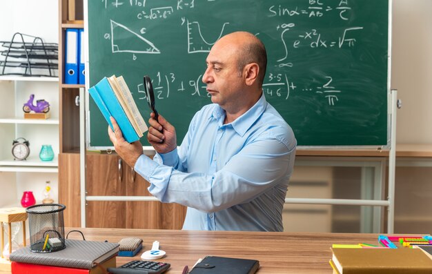 Pensando maestro de mediana edad se sienta a la mesa con útiles escolares libro de lectura con lupa en el aula