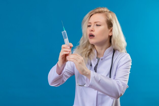 Pensando médico joven con estetoscopio en bata médica sosteniendo una jeringa sobre fondo azul.