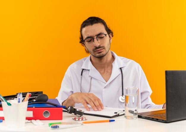 Pensando en el médico varón joven con gafas médicas vistiendo bata médica con estetoscopio sentados en el escritorio