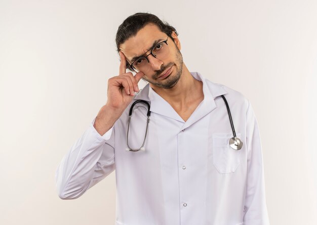Pensando en el médico varón joven con gafas ópticas vistiendo túnica blanca con estetoscopio poniendo la mano a la cabeza en blanco