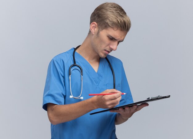 Pensando en el médico varón joven vistiendo uniforme médico con estetoscopio escribiendo algo en el portapapeles aislado en la pared blanca