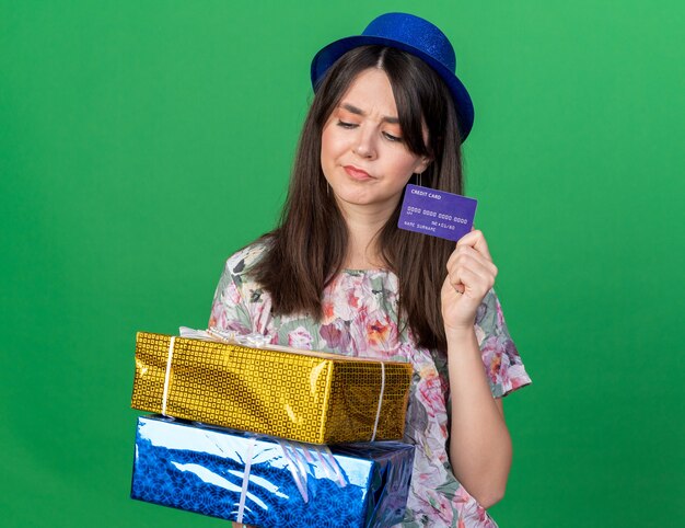 Pensando mirando hacia abajo hermosa joven vistiendo gorro de fiesta sosteniendo cajas de regalo con tarjeta de crédito aislado en la pared verde