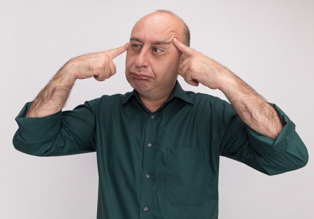 Pensando mirando al lado del hombre de mediana edad con camiseta verde poniendo los dedos en la sien aislado en la pared blanca