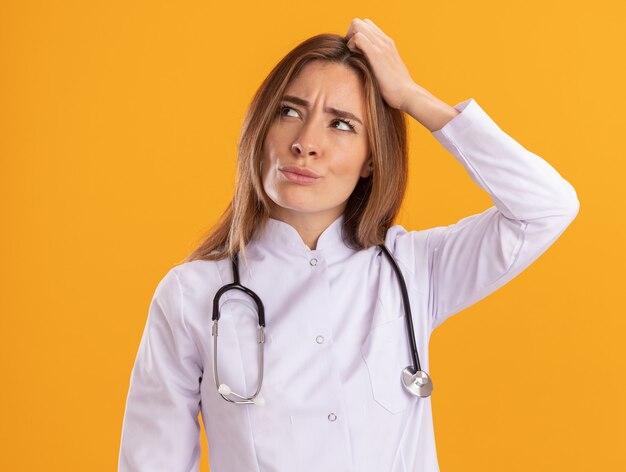 Pensando mirando al lado joven doctora vistiendo bata médica con estetoscopio rascándose la cabeza aislada en la pared amarilla