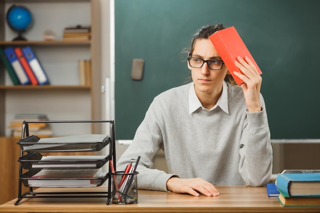 pensando mirando al lado joven maestro sosteniendo un libro en la cabeza sentado en el escritorio con herramientas escolares en el aula