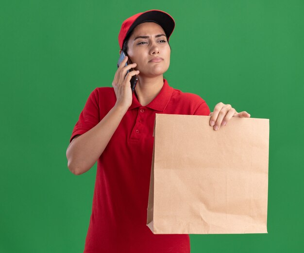 Pensando mirando al lado joven repartidora vestida con uniforme y gorra sosteniendo el paquete de comida de papel y habla por teléfono aislado en la pared verde