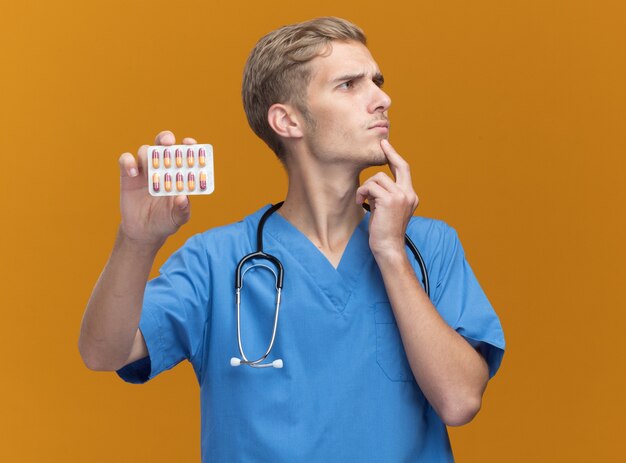 Pensando mirando al lado médico varón joven vistiendo uniforme médico con estetoscopio sosteniendo pastillas poniendo la mano debajo de la barbilla aislada en la pared naranja