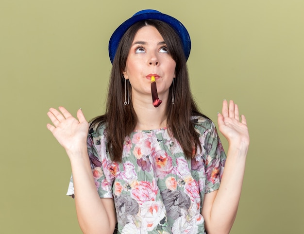 Pensando mirando hacia arriba joven hermosa mujer vistiendo gorro de fiesta soplando silbato de fiesta extendiendo las manos aisladas en la pared verde oliva