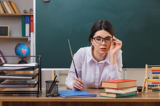 pensando mirando a la cámara joven maestra con gafas sosteniendo un puntero sentado en el escritorio con herramientas escolares en el aula