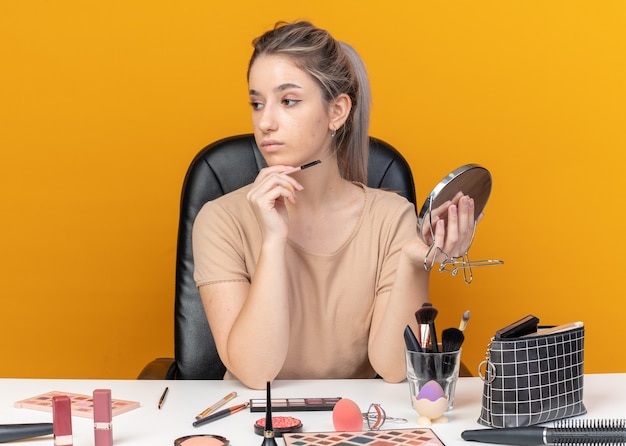 Pensando mirando de lado hermosa joven se sienta a la mesa con herramientas de maquillaje sosteniendo pincel de maquillaje con espejo aislado sobre fondo naranja