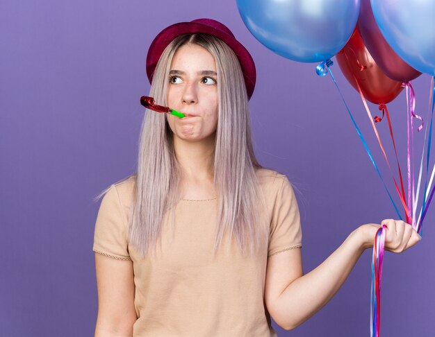 Pensando mirando de lado hermosa joven vistiendo gorro de fiesta sosteniendo globos soplando silbato de fiesta