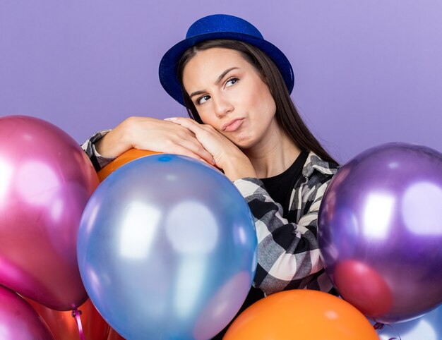 Pensando mirando hacia el lado joven hermosa mujer vistiendo gorro de fiesta de pie detrás de globos aislados en la pared azul