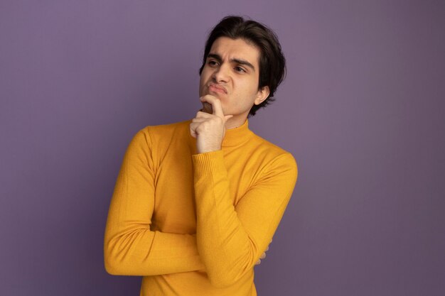 Pensando en mirar al lado chico guapo joven con suéter de cuello alto amarillo poniendo la mano en la barbilla aislada en la pared púrpura