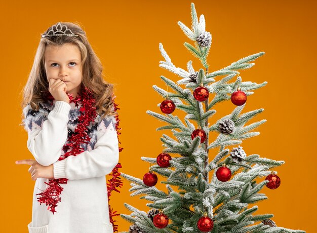 Pensando en mirar hacia arriba niña de pie cerca del árbol de Navidad con tiara con guirnalda en el cuello poniendo la mano en el mentón aislado sobre fondo naranja