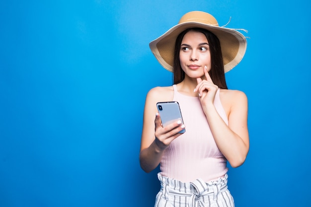 Pensando en la mujer joven con sombrero de paja mediante teléfono móvil aislado sobre la pared azul.