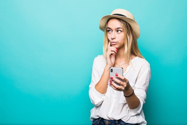 Pensando en la mujer joven con sombrero de paja mediante teléfono móvil aislado sobre pared azul