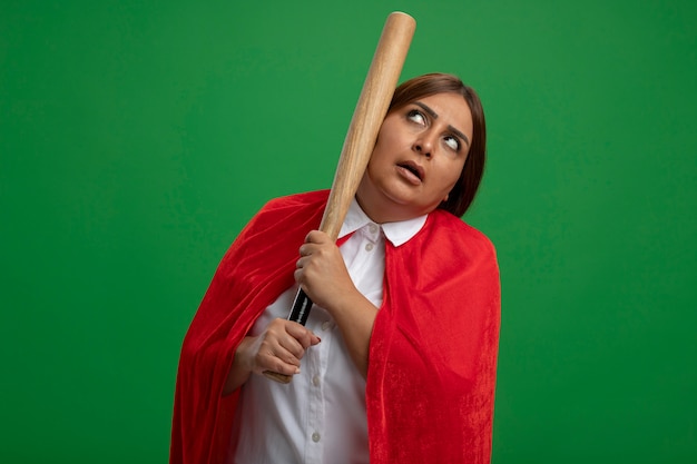 Pensando mujer de superhéroe de mediana edad mirando hacia arriba poniendo el bate de béisbol en la mejilla aislado sobre fondo verde