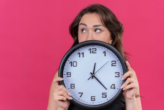 Pensando en niña caucásica vistiendo camiseta negra con reloj de pared sobre fondo rosa