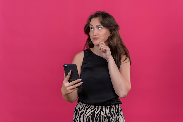 Pensando en niña caucásica vistiendo camiseta negra sosteniendo un teléfono sobre fondo rosa