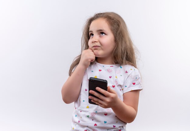 Pensando en la niña de la escuela con camiseta blanca sosteniendo el teléfono puso su mano en la mejilla en la pared blanca aislada