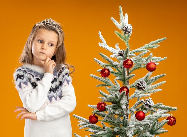 Pensando niña de pie cerca del árbol de Navidad con tiara con guirnalda en el cuello poniendo la mano debajo de la barbilla aislado en la pared naranja