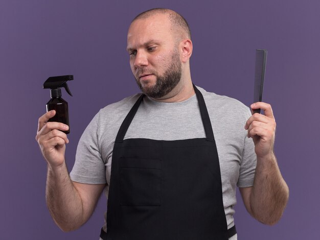 Pensando en el peluquero masculino de mediana edad eslavo en uniforme sosteniendo el peine y mirando la botella de spray en la mano aislada en la pared púrpura