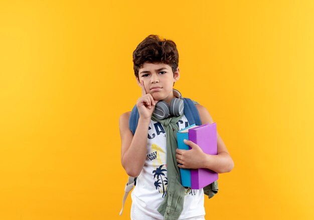 Pensando en el pequeño niño de la escuela con mochila y auriculares sosteniendo libros y poniendo la mano en la mejilla aislada en amarillo