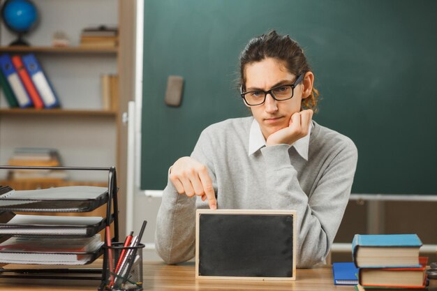 pensando en poner la mano en la barbilla joven maestro con anteojos sentado en el escritorio sosteniendo una mini pizarra con herramientas escolares en el aula