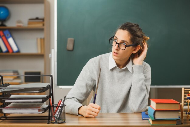 Pensando en poner la mano en la cabeza, un joven maestro con gafas sosteniendo un puntero sentado en el escritorio con herramientas escolares en el aula