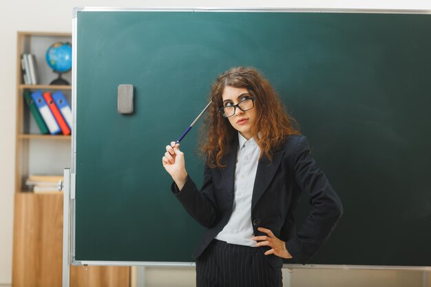 pensando en poner la mano en la cadera joven maestra sosteniendo un puntero de pie frente a la pizarra en el aula