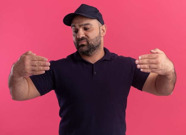 Pensando en el repartidor de mediana edad en uniforme y gorra fingiendo sostener algo aislado en la pared rosa