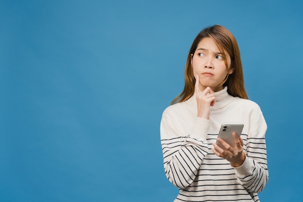 Pensando soñando a la joven dama de Asia usando el teléfono con expresión positiva, vestida con ropa casual sintiendo felicidad y parada aislada en la pared azul