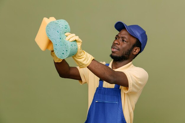 Pensando en sostener esponjas de limpieza joven limpiador afroamericano en uniforme con guantes aislados en fondo verde
